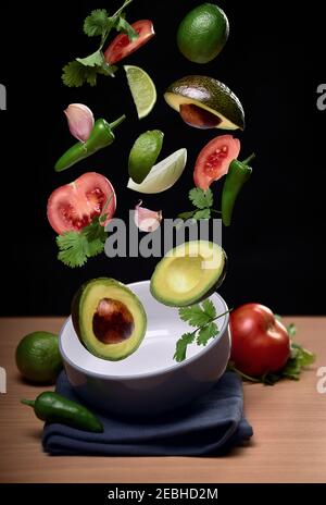 Guacamole Zutaten fliegen über Schüssel auf schwarzem festen Hintergrund. Tomaten, Pfeffer, Zwiebeln, Knoblauch, Avocado, Koriander, Limette Stockfoto
