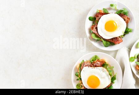 Offene Sandwiches mit Spiegeleiern, Speck und Gemüseblättern auf dem Teller auf hellem Steinhintergrund mit Freitextbereich. Draufsicht, flach liegend Stockfoto