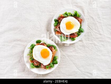 Offene Sandwiches mit Spiegeleiern, Speck und Gemüseblättern auf dem Teller auf hellem Hintergrund. Draufsicht, flach liegend Stockfoto