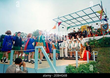 Europareise: Irland, Galway: Präsident Kennedy am Eyre Square, Bemerkungen und Abreise, 11:55am Uhr. Präsident John F. Kennedy (oben rechts), Bürgermeister von Galway, Alderman Patrick D. Ryan (links von Präsident Kennedy), Und andere hochrangige Gäste beobachten von der oberen Plattform aus, wie Kinder von lokalen Schulen (unten links) während einer Begrüßungszeremonie für den Präsidenten auf dem Eyre Square in Galway, Irland, einen traditionellen irischen Tanz vorführen; Kinder des Patrizier Brothers Choir (rechts von den Tänzern) singen, begleitet von der Harfenistin Ruth Bradley (sitzend, mit rotem Rock). Auch abgebildet (sitzt in der Gruppe Stockfoto