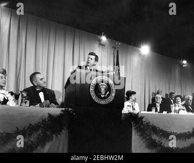 National Cultural Center Fonds-Raising Benefit, 9:23pm. Präsident John F. Kennedy (am Rednerpult) hält Bemerkungen bei einem Fundraising Dinner und Closed-Circuit Telecast von u0022An American Pageant der Artsu0022 für das National Cultural Center. Seated (L-R): vorsitzender des Dinner Committee für das National Cultural Center, Suzanne Gardner; Vice President Lyndon B. Johnson; First Lady Jacqueline Kennedy; Vorsitzender des Kuratoriums für das National Cultural Center, Roger L. Stevens; Lady Bird Johnson; und Chief Justice des Supreme Court, Earl Warren. National Guard Armory, Washington, Stockfoto