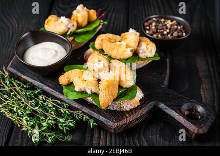 Toasts mit knusprig gebratenen Garnelen und grünem Salat auf einem Holzbrett. Schwarzer Holzhintergrund. Draufsicht Stockfoto