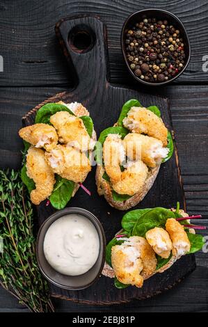 Toasts mit knusprig gebratenen Garnelen und grünem Salat auf einem Holzbrett. Schwarzer Holzhintergrund. Draufsicht Stockfoto