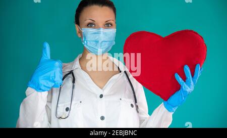 Eine Kardiologin hält ihren Daumen hoch und ein großes rotes Herz. Ein gesundes Herz in den Händen eines Arztes. Stockfoto
