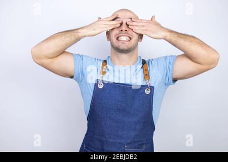 Junge Glatze Mann trägt Schürze Uniform über isolierten weißen Hintergrund Augen mit Händen lächelnd fröhlich und lustig. Blindes Konzept. Stockfoto