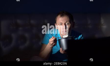 Ein junger Mann schaut nachts Fernsehen und isst Eis. Überraschendes Gesicht Stockfoto