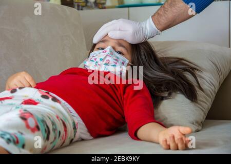 Handschuhen männliche Hand misst die Temperatur des Mädchens tragen Eine schützende Gesichtsmaske Stockfoto