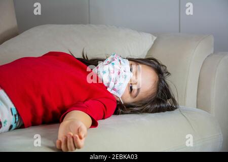 Kleine kranke Mädchen trägt schützende Gesichtsmaske liegt auf dem Sofa Stockfoto