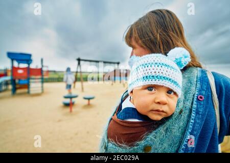 Eine junge Mutter steht mit ihr auf einem Spielplatz Baby in einem Tragetuch Stockfoto