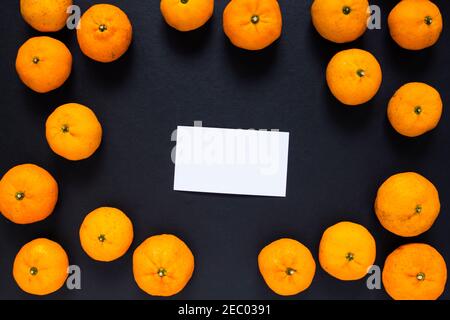 Leere Visitenkarte und orangefarbene Mandarinen auf schwarzem Hintergrund. Saftige Orangen Foto Hintergrund. Weiße Papierkarte mit Textplatz. Blick von oben auf Zitrusfrüchte. Stockfoto