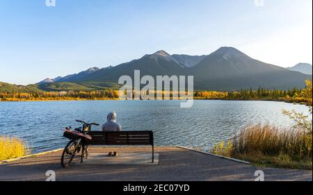 Touristen ruhen auf Holzbank, entspannen am Vermilion Lakes Seeufer im Herbst Laubsaison Sonnenuntergang Zeit. Radfahren im Banff National Park Stockfoto