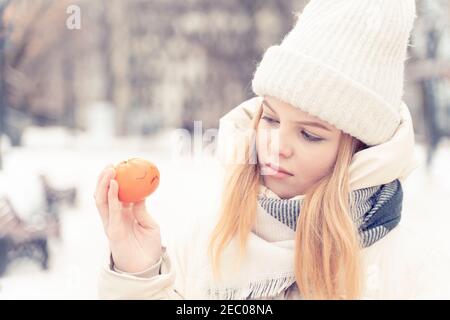 Schöne kaukasische Mädchen Teenager in einem weißen Hut und Daunenjacke einsam im Winter. Eine junge Frau spricht mit einer Mandarine, auf die sie eine sa gemalt Stockfoto