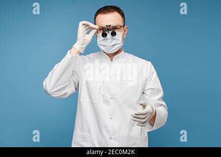 Kaukasischer Arzt in einer OP-Maske, Schutzhandschuhe und binokulare Lupen auf dem blauen Hintergrund, isoliert mit Kopierraum für Text oder Produkt Stockfoto