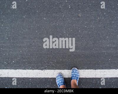 Grauer Asphalt mit weißer Linienmarkierung und Frauenfüßen in Schuhen. Mädchen Füße in Komfort-Schuhe überqueren weiße Linie auf Asphalt. Straßenmarkierung, Ansicht von oben. Verkehrsregeln Stockfoto