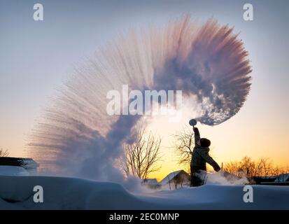 Der Kerl spritzt gekochtes Wasser aus einer Thermoskanne über seinen Kopf vor dem Hintergrund eines Sonnenuntergangs bei frostigen Wetter. Die Wirkung des sofortigen Einfrierens von heißem w Stockfoto