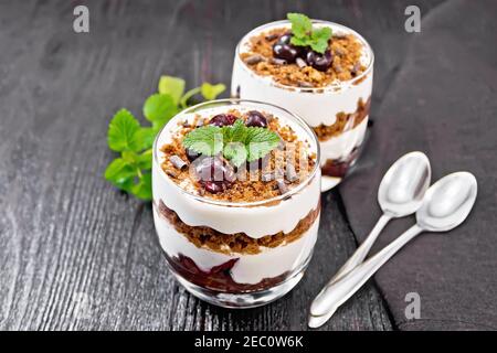 Dessert Schwarzwald von Kirschen, Schokoladenkeks und weichen Quark mit Sahne in zwei Gläsern, ein Handtuch, Minze und ein Löffel auf dunklem Holzbrett Stockfoto