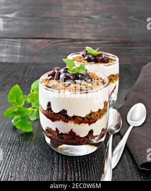 Dessert Schwarzwald mit Kirschen, Schokoladenkeks und weichem Hüttenkäse mit Sahne in zwei Gläsern, Serviette, Minze und Löffel auf dunklem Holzbrett Stockfoto