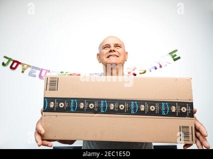 Paris, Frankreich - Oct 10, 2019: Lächelnd glücklich Senior Mann lächelnd, während er große neue Amazon Prime Paket-Logotyp mit dem lächelnden Pfeil hält - Happy Birthday Signage an der Wand im Hintergrund Stockfoto