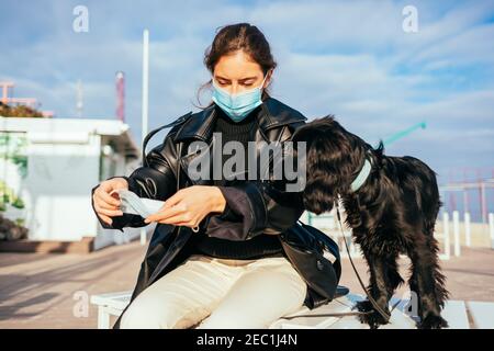 Junge Frau mit medizinischer Maske sitzt im Freien neben ihrem Hund mit einer anderen Maske für ihr Haustier. Stockfoto