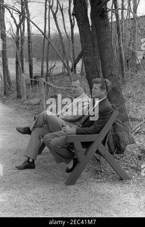 Wochenende im Camp David. Präsident John F. Kennedy (rechts, hält Pfeife) sitzt auf einer Bank mit Kennedy Familie Freund und Untersekretär der Marine, Paul u0022Redu0022 Fay, in Camp David in Frederick County, Maryland. Stockfoto