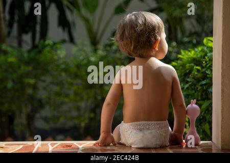 Ein charmantes Kleinkind sitzt mit einem Spielzeug auf den Stufen eines Hauses im Sonnenlicht. Rückansicht. Stockfoto