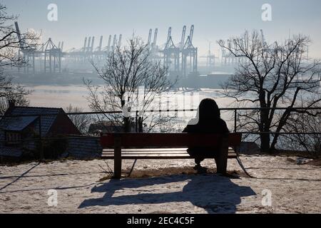 Hamburg, Deutschland. Februar 2021, 13th. Eine Person sitzt auf einer Bank und genießt die Sonne und den Blick auf die teilweise gefrorene Elbe und die Kraniche im Hamburger Hafen. Quelle: Georg Wendt/dpa/Alamy Live News Stockfoto