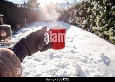 Person hält eine rote Tasse mit heißem Kaffee, Tee oder Schokoladenmilch mit dampfenden Rauch und Handschuhe im Schnee, Winter, Entspannung, trinken, Schnee, gemütliches Konzept Stockfoto