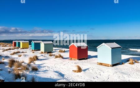 FINDHORN BAY MORAY FIRTH SCHOTTLAND BLAUER HIMMEL UND SIEBEN BUNTE HÜTTEN ODER CHALETS AN EINEM SCHNEEBEDECKTEN STRAND IM WINTER Stockfoto