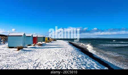 FINDHORN BAY MORAY FIRTH SCHOTTLAND GROSSE WELLEN AM MEER UND SEHR BUNTE HÜTTEN ODER CHALETS AN EINEM SCHNEEBEDECKTEN STRAND IM WINTER Stockfoto