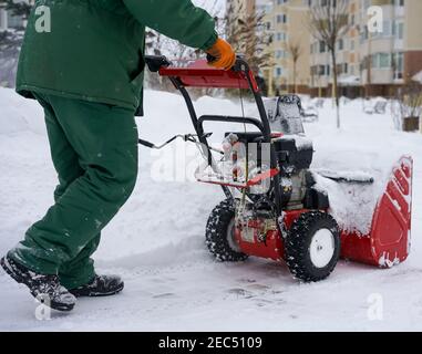 Ein Arbeiter in grünen Overalls entfernt Schnee mit einem roten Schneefräsen vor dem Hintergrund einer Fichte, einer Schneeverwehung und einem Haus. Stockfoto