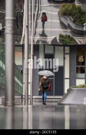 Eine Frau, die während der Ausgießung in der Stadtstraße wegging Heftiger Regen mit Regenschirm bis ihre Reflexion in Metall Chrom Gebäudeoberflächenwand darüber Stockfoto