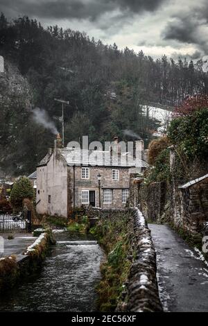 Castleton Hope Valley ländliche Dorfhütte mit Rauch aus Kamin Peak District Fluss fließt im Winter mit Schnee und dramatisch Sturm Wolken Himmel Stockfoto