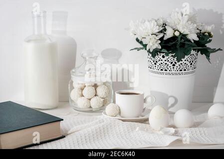 Flasche Milch, Buch, Kokos-Bonbons im Glas, Tasse Espresso-Kaffee, Blumentopf mit chrysanthen, Eier, Küchentuch auf dem Tisch. Stockfoto