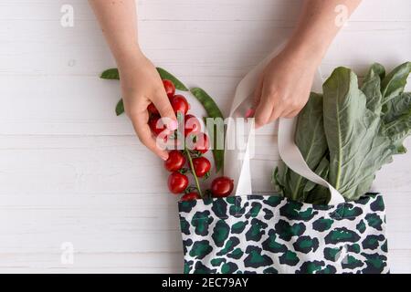 Hände einer Frau nehmen frische Bio-Tomaten aus dem Eco Shopping Bag mit trendigem Leopardenmuster-Print auf Holzhintergrund, Flat Lay. Draufsicht. Eco Stockfoto