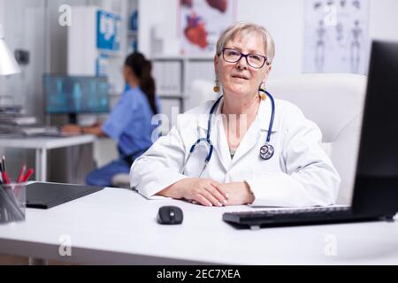 Selbstbewusster erfahrener Senior-Arzt im weißen Mantel mit Stethoskop, das Videokonferenz- und Beratungspartner oder Patienten hält, Blick in die Kamera. Arzt informiert den Patienten über einen Videoanruf Stockfoto