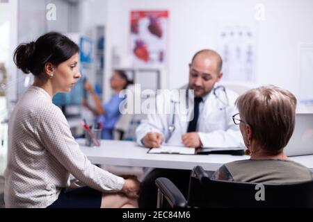 Doughter und behinderte Mutter im Rollstuhl sitzend im Gespräch mit dem Arzt während der Konsultation. Praktiker Fragen stellen, um Patienten füllen Krankheitsgeschichte Form reifen, verschreiben Behandlung Stockfoto
