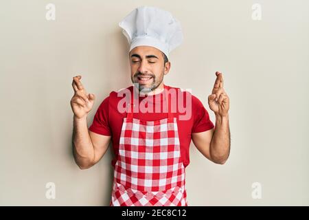 Junge hispanic Mann trägt Bäcker Uniform gestikulierenden Finger überquert lächelnd mit Hoffnung und Augen geschlossen. Glück und abergläubische Konzept. Stockfoto