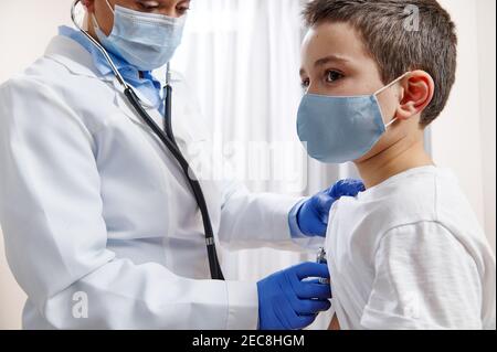 Kinderärztin in der medizinischen Uniform verwendet ein Stethoskop beim Auskultieren Ein kleiner Junge in schützender medizinischer Maske Stockfoto