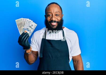 Junger afroamerikanischer Mann, der professionelle Schürze mit USA-Dollar trägt Banknoten, die positiv und glücklich stehen und lächeln mit einem Selbstbewusste s Stockfoto