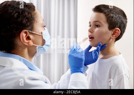 Afroamerikanischer Arzt mit Gesichtsmaske und Schutzhandschuhen, die bei einem Hausbesuch die Kehle eines Jungen untersuchen. Stockfoto