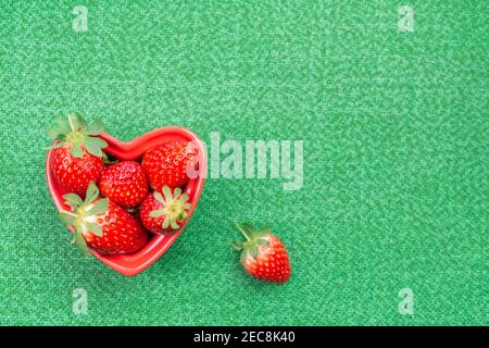 Rote frische und reife Erdbeeren in roter herzförmiger Schale auf Tisch, überzogene grüne Tischdecke, Hintergrund. Draufsicht, flach liegend, Kopierbereich. Liebe und Valen Stockfoto