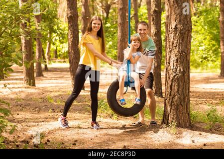 Portrait von jungen glücklichen Eltern, die Tochter auf Rad schwingen, mit Sommer Natur auf Hintergrund Stockfoto