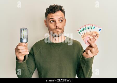 Junger hispanischer Mann hält LED-Birne und Euro-Banknoten lächelnd Blick zur Seite und starrt weg denken. Stockfoto