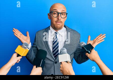 Kahlköpfiger Mann mit Bart, der von Reportern mit Mikrofonen ohne Ahnung und verwirrt mit offenen Armen interviewt wurde, keine Ahnung und zweifelhaftes Gesicht. Stockfoto