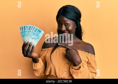 Afrikanische junge Frau trägt Arzt Uniform hält 100 brasilianischen reals Lächelnd glücklich zeigen mit Hand und Finger Stockfoto