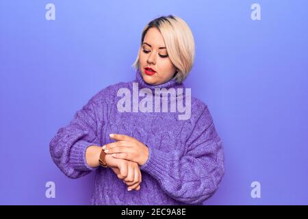 Schöne blonde plus size Frau trägt lässige Rollkragenpullover über lila Hintergrund Überprüfung der Zeit auf Armbanduhr, entspannt und zuversichtlich Stockfoto