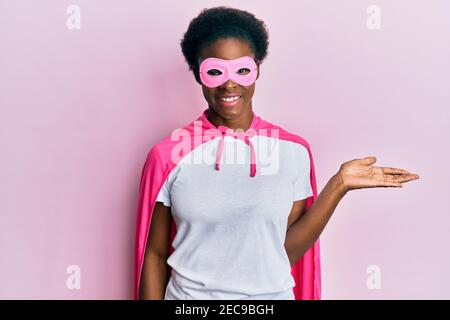 Junge afroamerikanische Mädchen trägt Superhelden Maske und Cape Kostüm lächelnd fröhlich präsentiert und zeigt mit Handfläche auf die Kamera zu schauen. Stockfoto