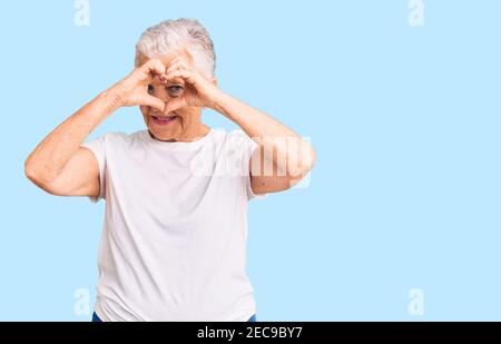 Ältere schöne Frau mit blauen Augen und grauen Haaren tragen Casual weißes T-Shirt tun Herzform mit Hand und Finger Lächelnd durch das Schild schauen Stockfoto