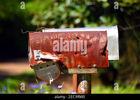 Verlassene Mailbox hat mehrere Einschusslöcher darin. Es ist rostig, verbogen und die Tür hängt kaum an. Eine neuere Mailbox befindet sich dahinter. Stockfoto