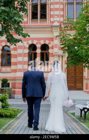 Vertikale Aufnahme eines frisch verheirateten Paares zu Fuß in einem park mit einem Gebäude im Hintergrund Stockfoto